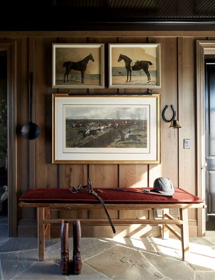 A wood-panelled foyer with equestrian prints, a velvet bench, and tall riding boots